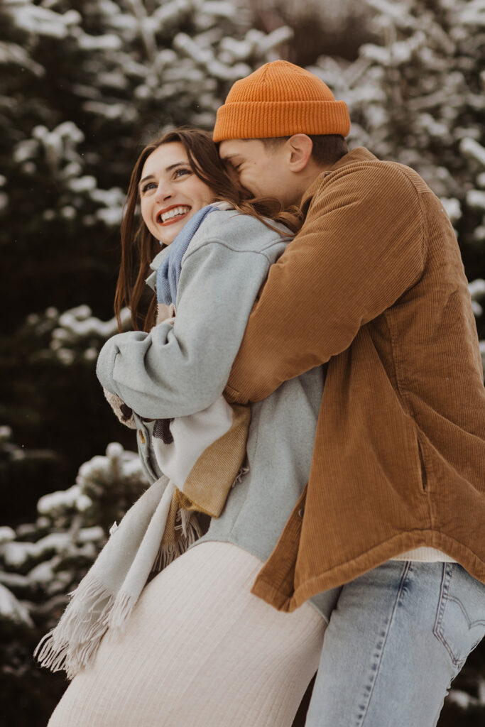 Man wearing camel jacket and orange hat nestles his face in his fiance's hair as he twirls her around