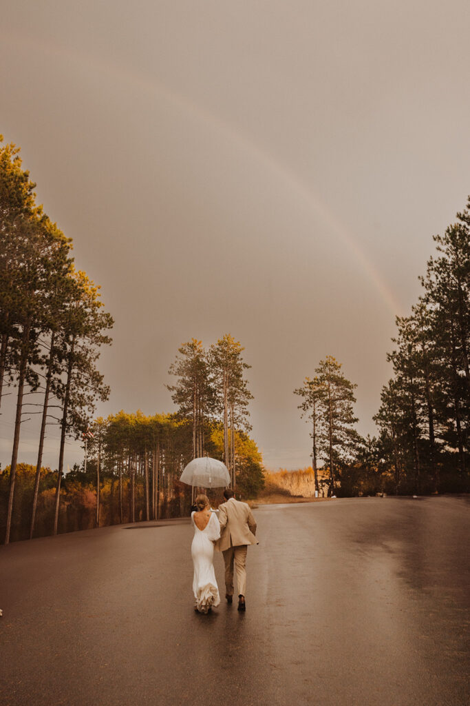 Bride and groom skip off into the distance while rainbow beams in sky above them