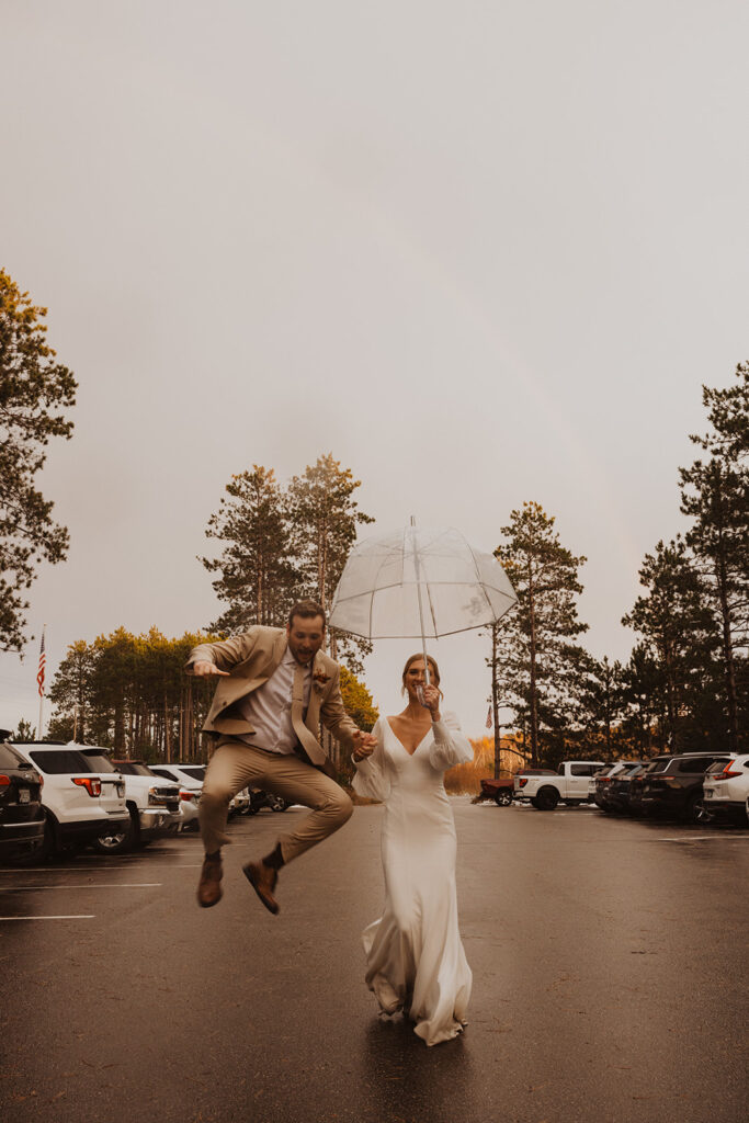 Groom leaps up into the air in excitement as bride trails behind holding umbrella