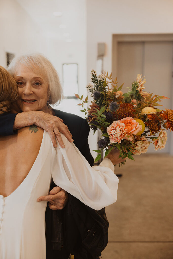 Grandmother hugs bride as she holds onto her colorful arrangement of winter wedding blooms