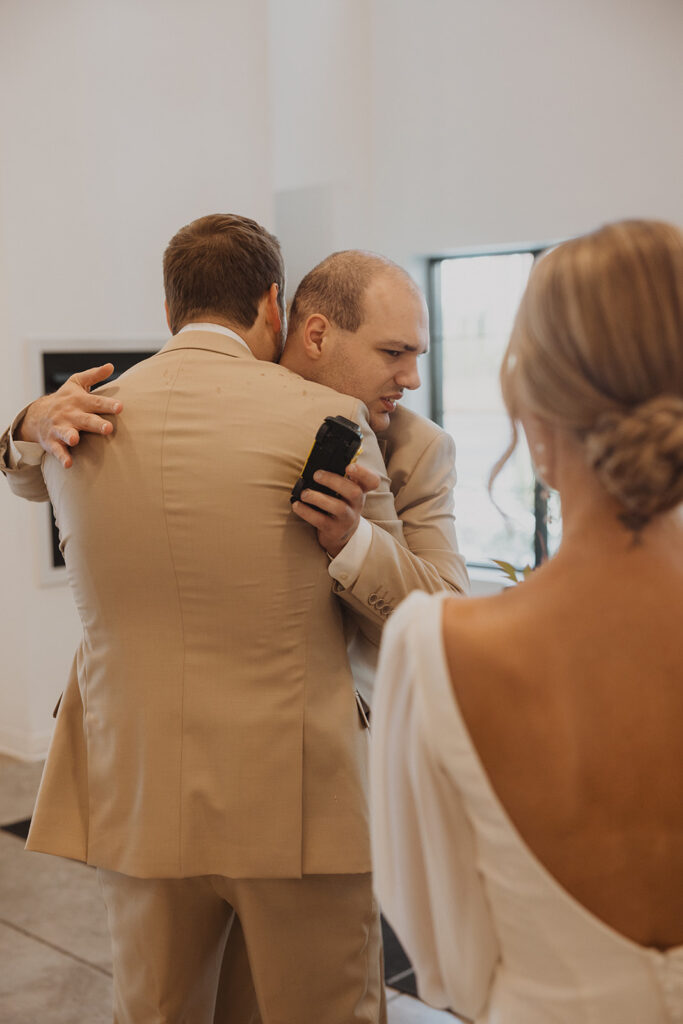 Groom hugs his brother in emotional embrace while bride looks on 