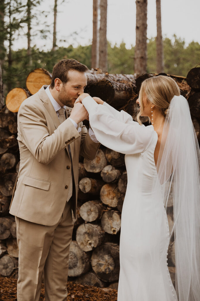 Groom kisses both of bride's hands near woodstack at Pinewood Weddings and Events