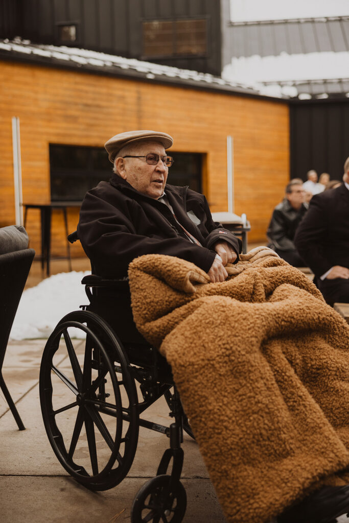 Elderly man watches granddaughter's winter wedding venue ceremony while bundled under a cozy blanket