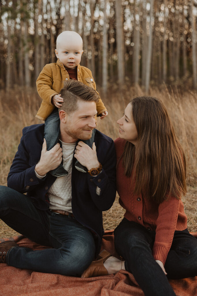 Mom and dad look at each other knowingly as baby sits on dad's shoulders