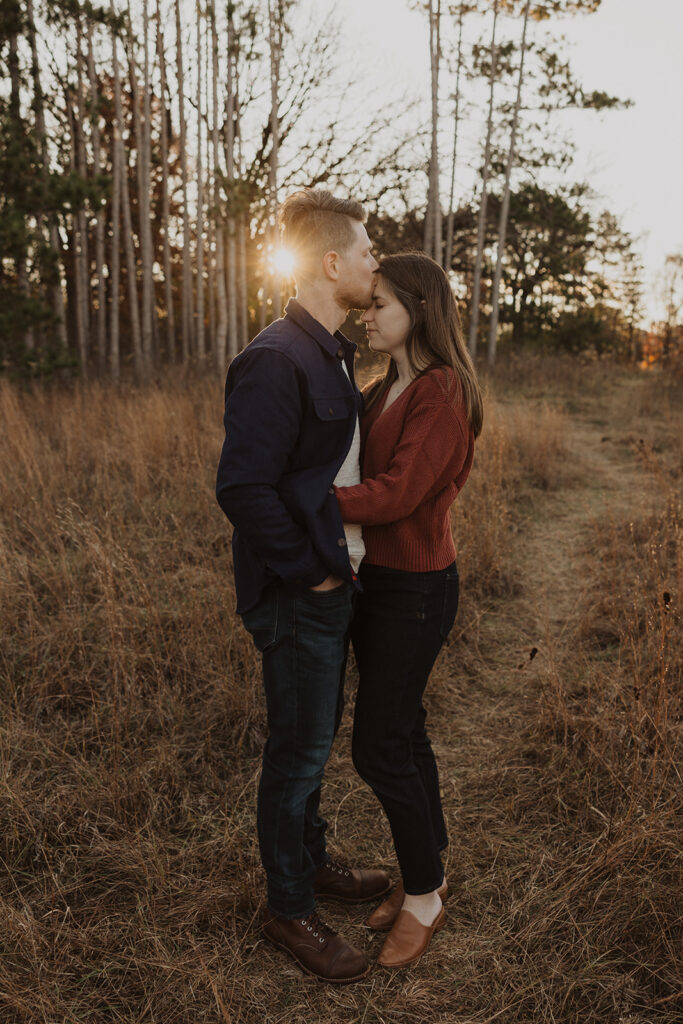 Husband kisses wife's forehead as the sun rises behind them during family photography with dogs