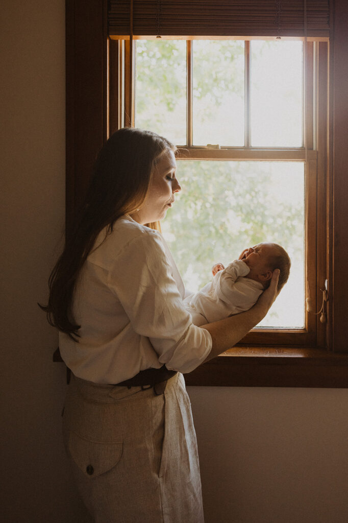 Mom soothes newborn baby in glow of window daylight in newborn photography with family