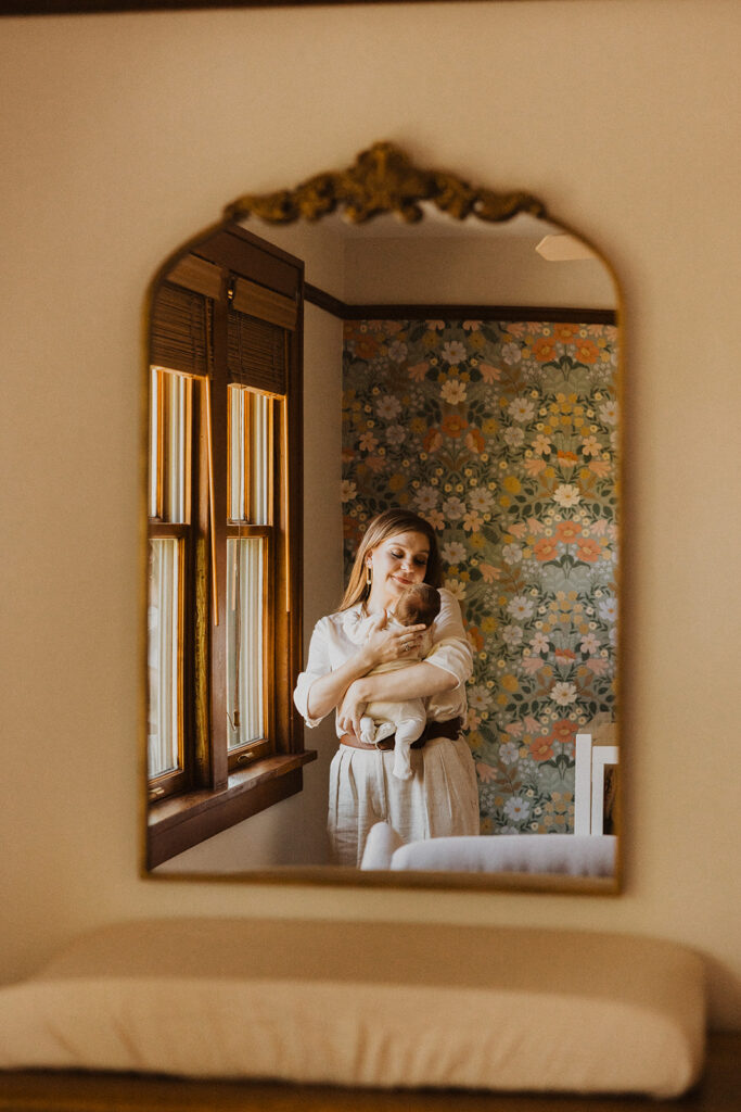 Mom rocks her newborn baby to sleep in reflection of gold vintage mirror