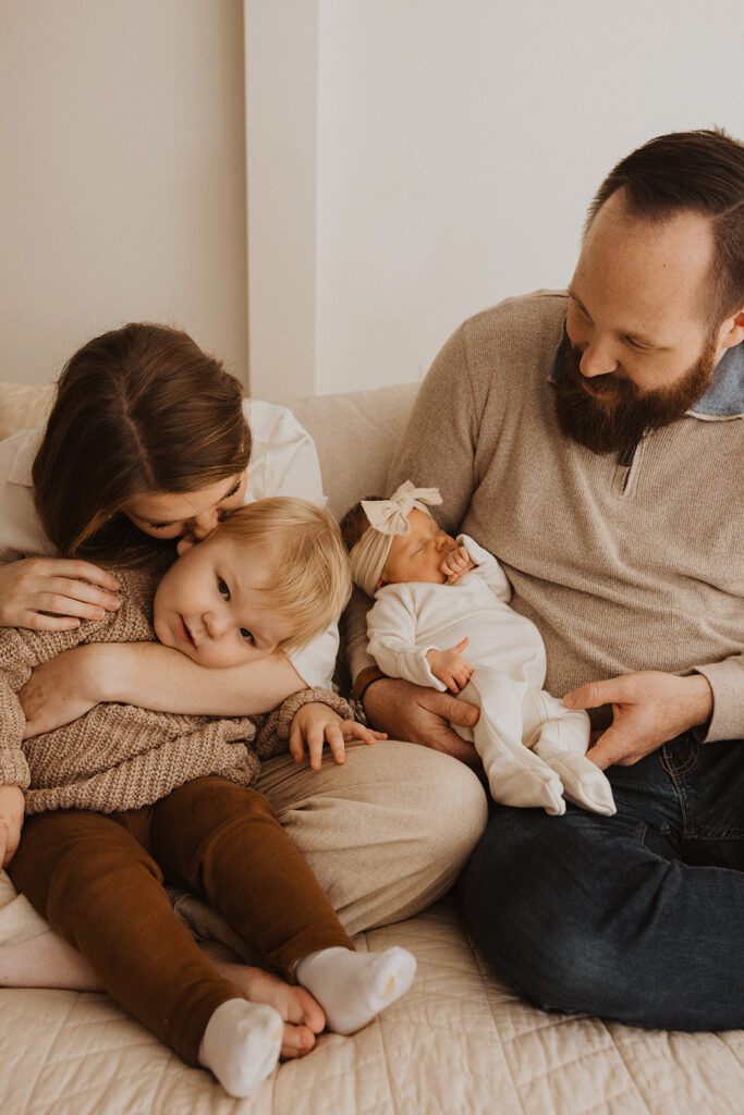 Mom kisses big brother's head while dad holds newborn baby in newborn photography with family