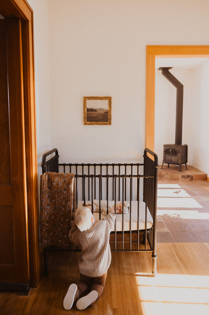 Toddler pokes his hands through the crib bars to touch his newborn baby sister