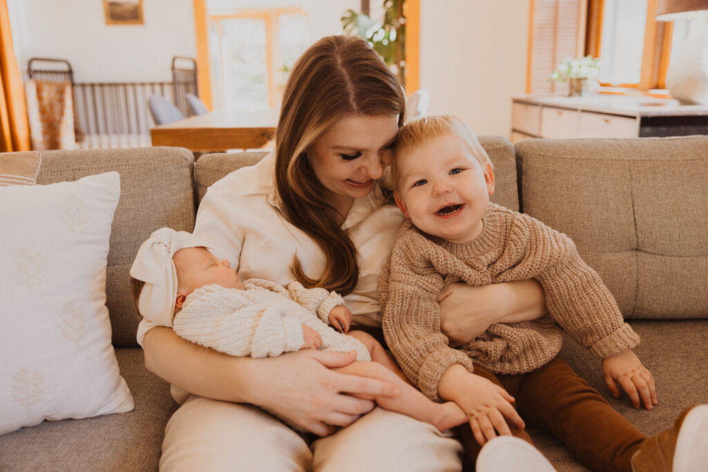 Mom and son sit joyously on beige couch holding newborn baby