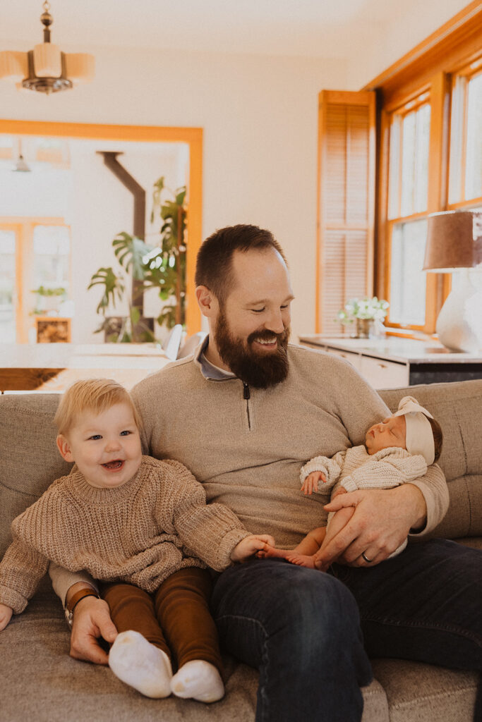 Dad and toddler son sit on couch with newborn baby