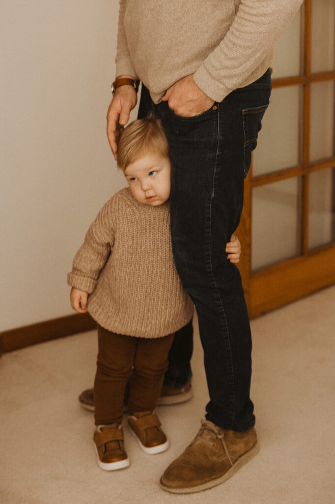 Young boy wraps his arms around his dad's leg as he watches mom change baby's diaper